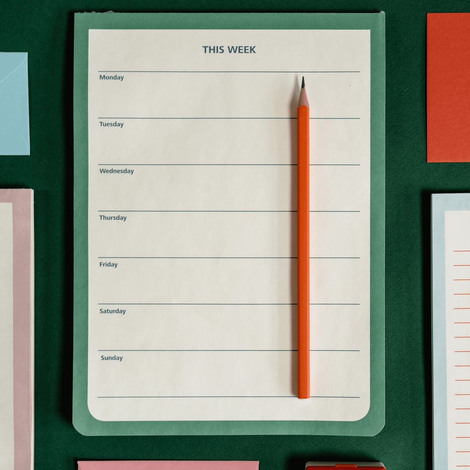 Weekly planner on green surface with an orange pencil. Days of the week visible. Colorful papers around create an organized mood.