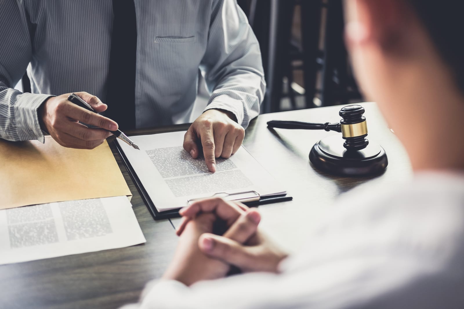 Two people in a meeting; one points at a document on a clipboard. A gavel is on the table. Formal setting, serious discussion.
