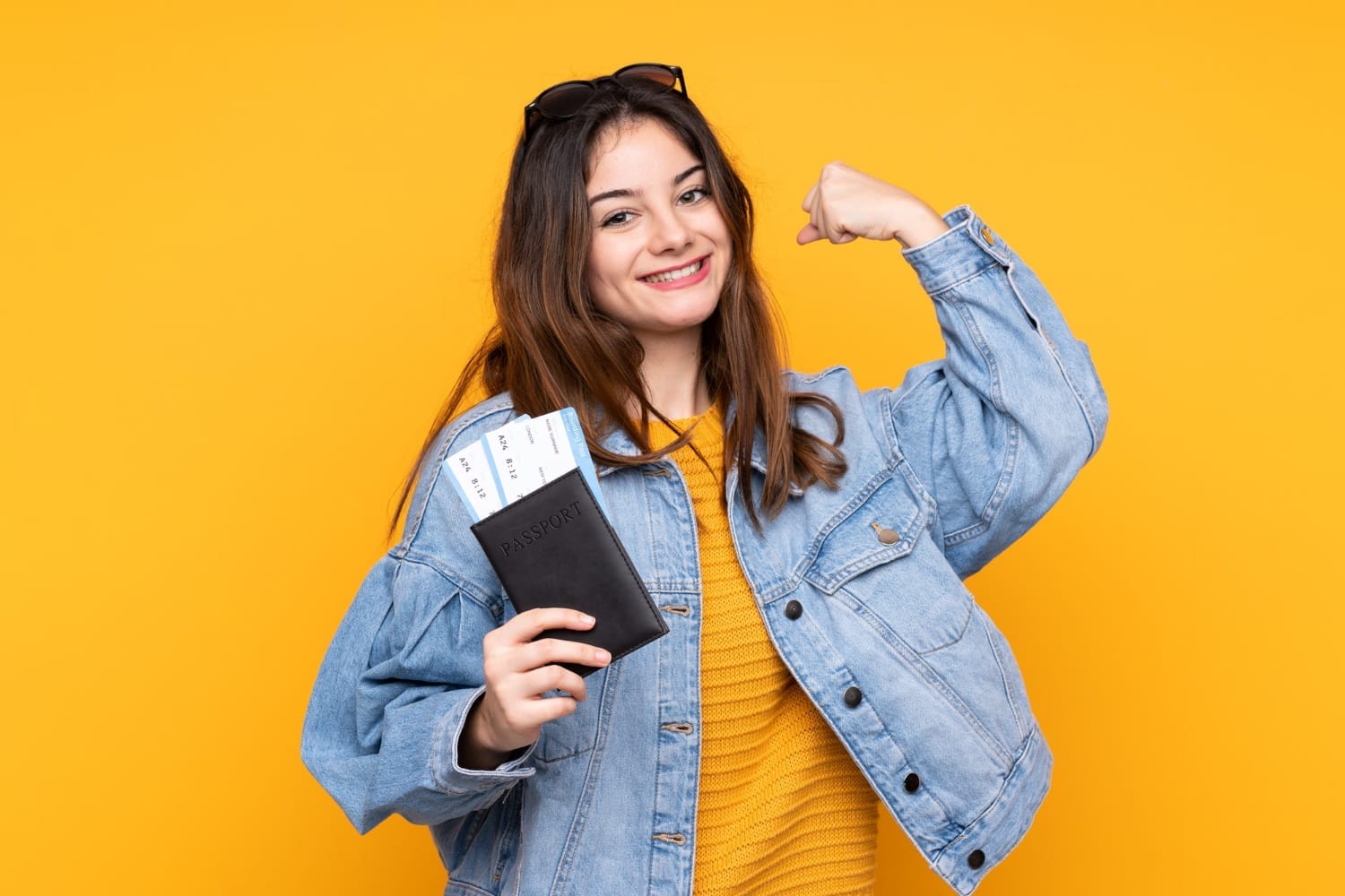 Smiling woman in a denim jacket holds a passport with tickets, flexing her arm against a bright yellow background. Mood is cheerful and excited.